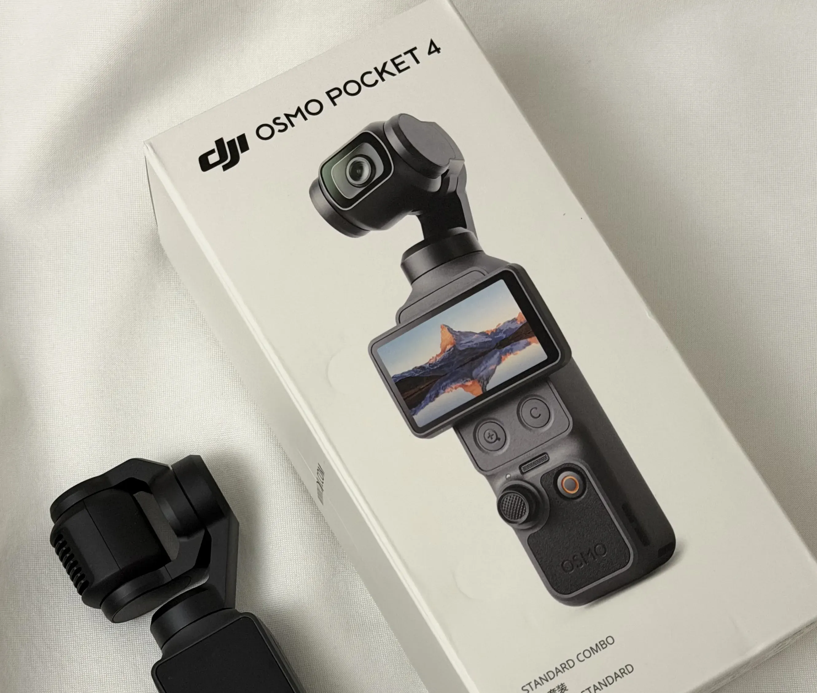 DJI Osmo Pocket 4 gimbal camera lying next to its white Standard Combo retail box on white fabric