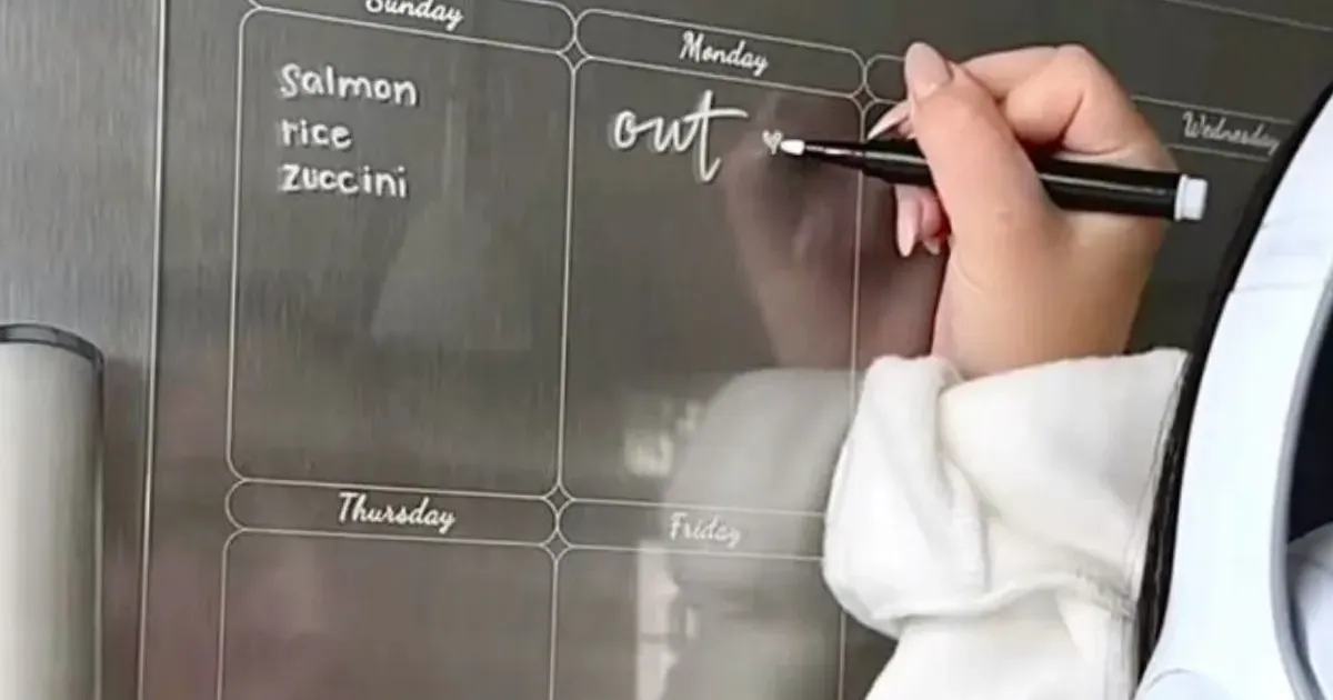 Hand schreibt den Wochenplan auf eine durchsichtige Acryl-Tafel an der Kühlschranktür