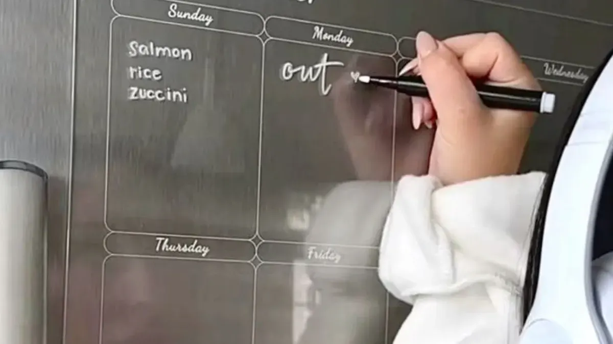 Hand writing the weekly menu on a clear acrylic planner board mounted on a fridge door