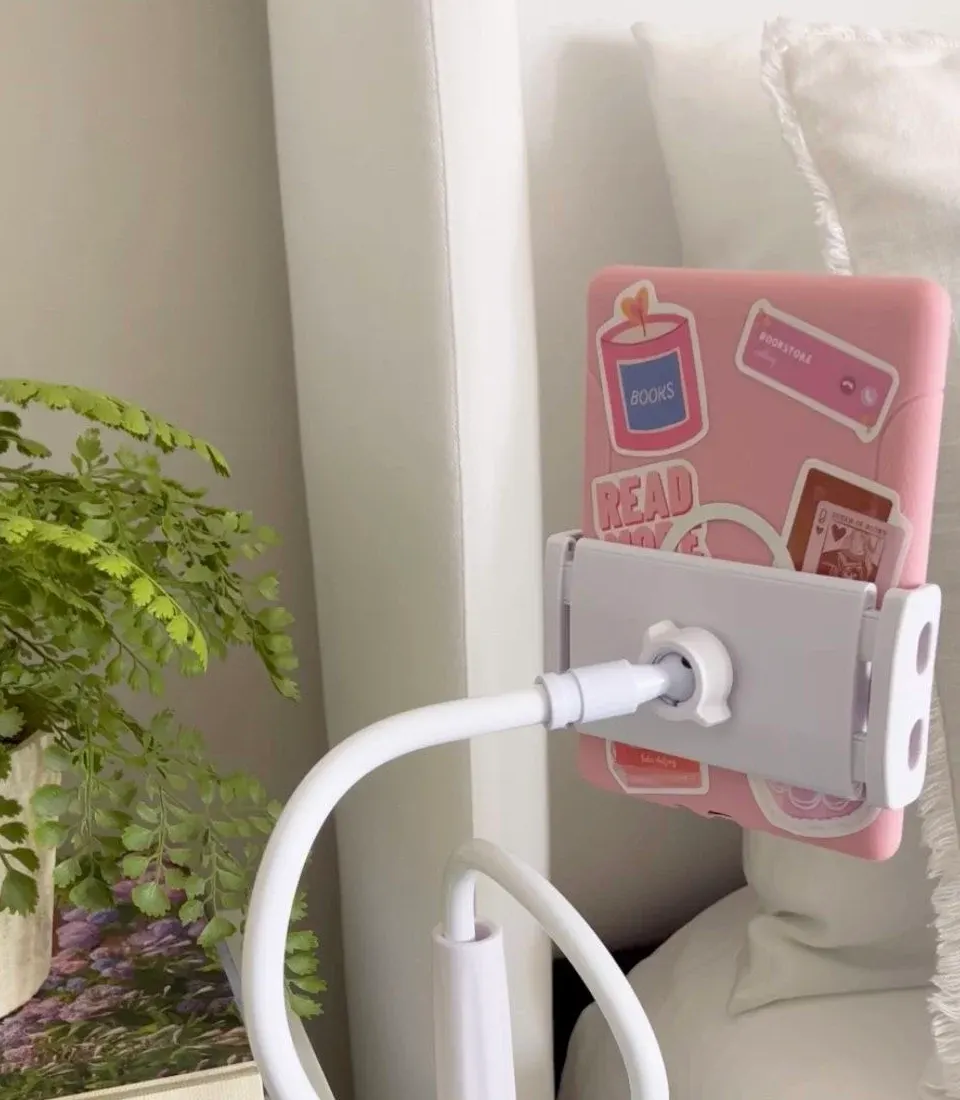 White gooseneck tablet holder clamped to a bedside table, holding a pink tablet with stickers above a bed