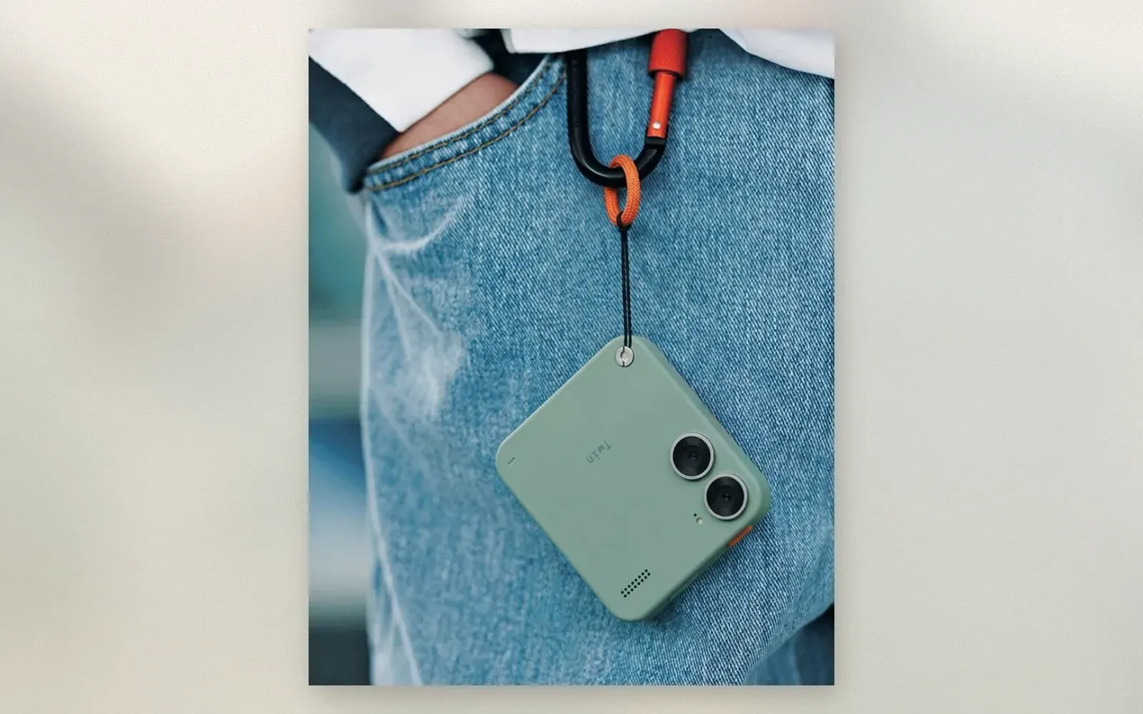 Small sage-green dual-lens mini camera hanging from a carabiner on jeans