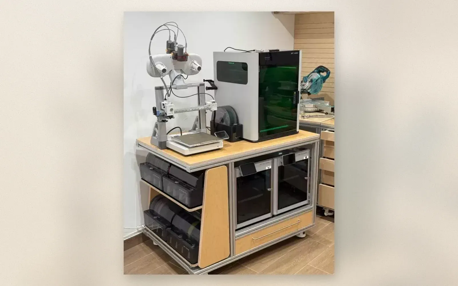Rolling plywood and aluminum workstation holding four 3D printers, with filament dry boxes on the side shelves