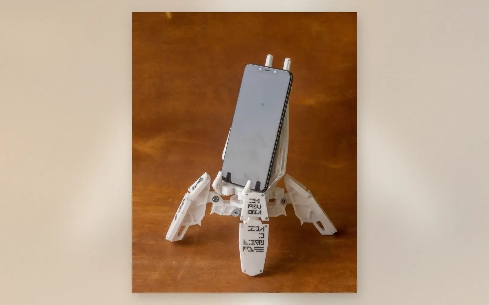 White 3D-printed mech exo-suit phone holder with articulated legs gripping a smartphone on a wooden desk