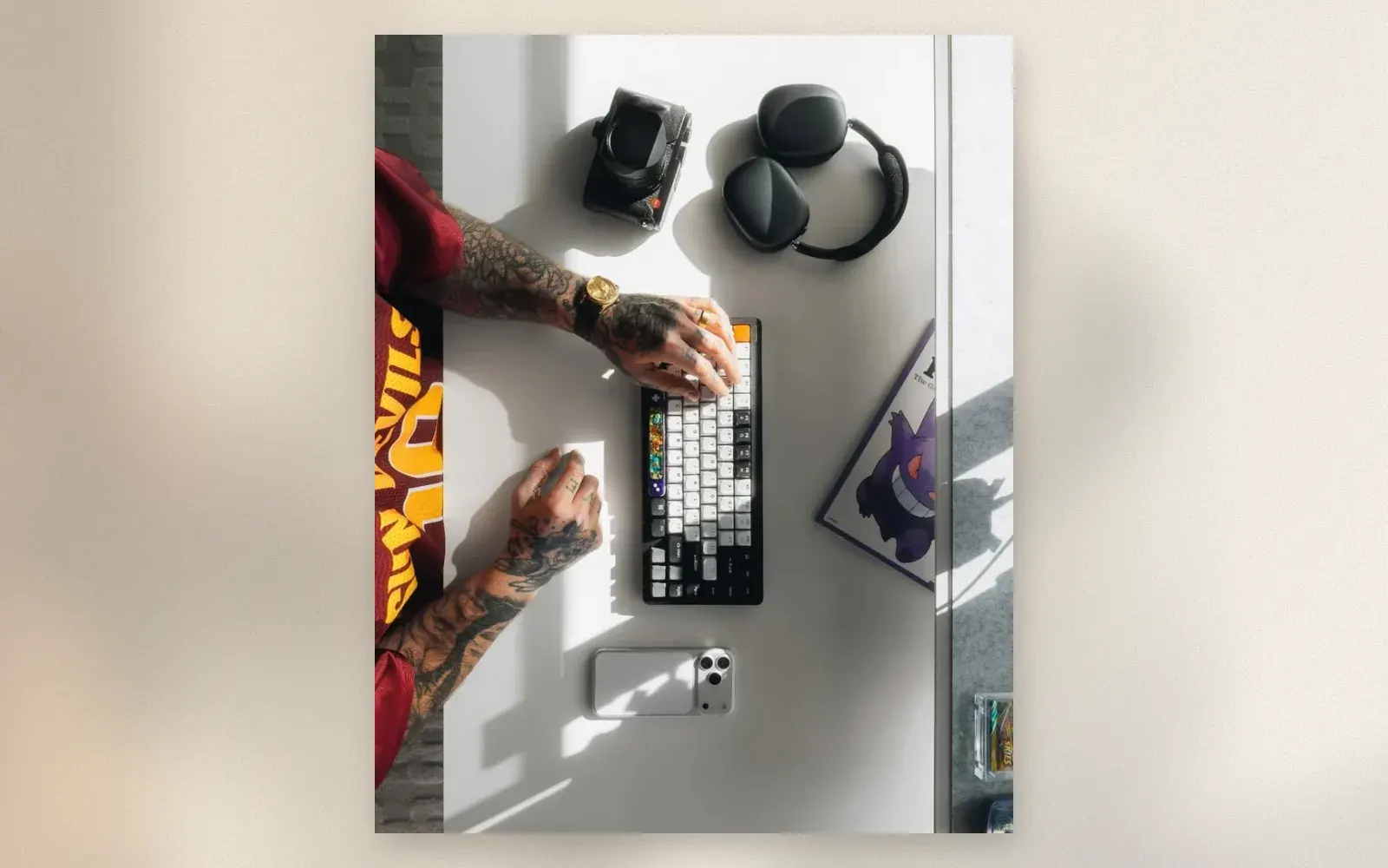 Top-down view of white desk with custom mechanical keyboard, over-ear headphones, compact camera, and iPhone