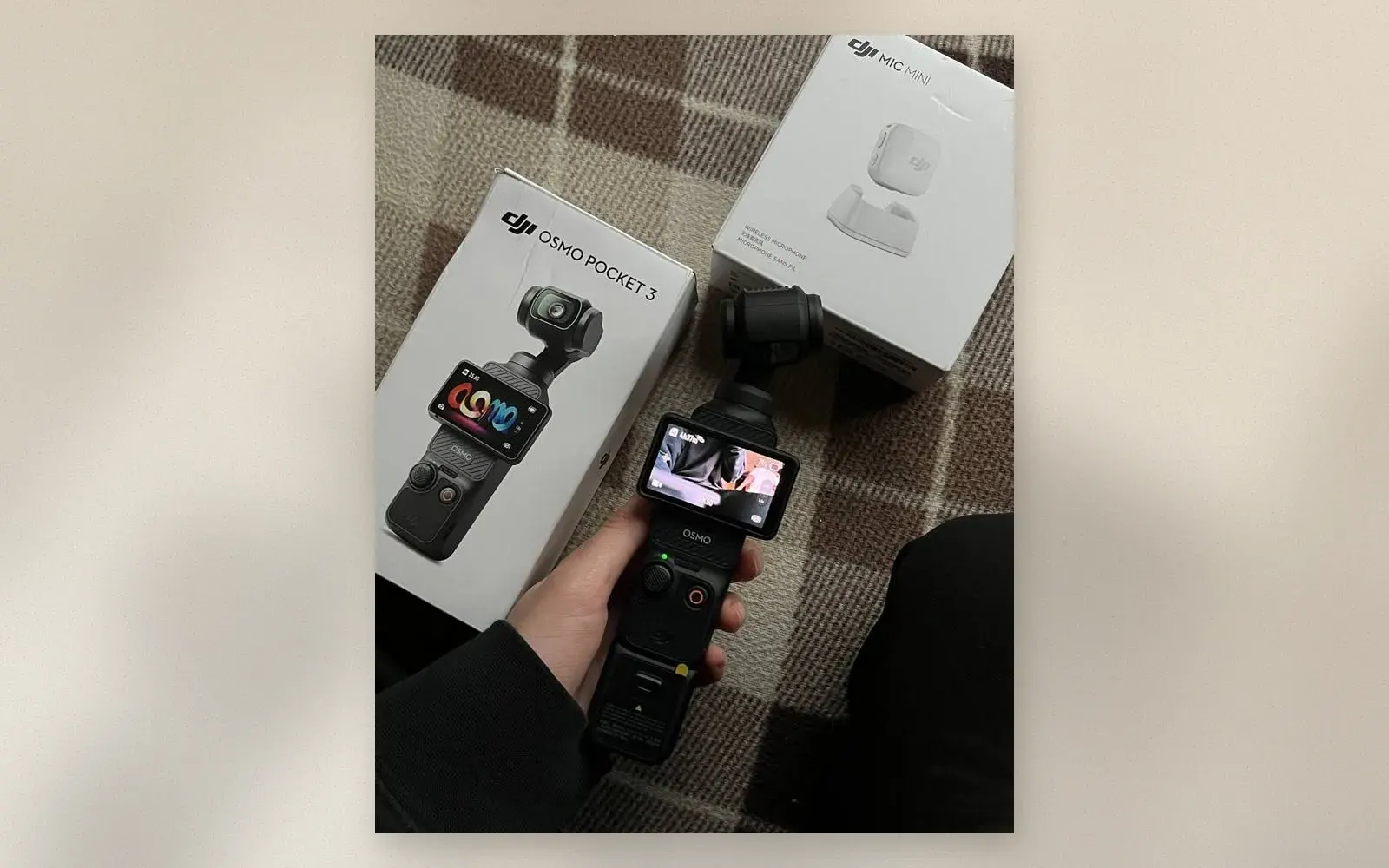 Hand holding the DJI Osmo Pocket 3 in front of its retail box and a DJI Mic Mini box