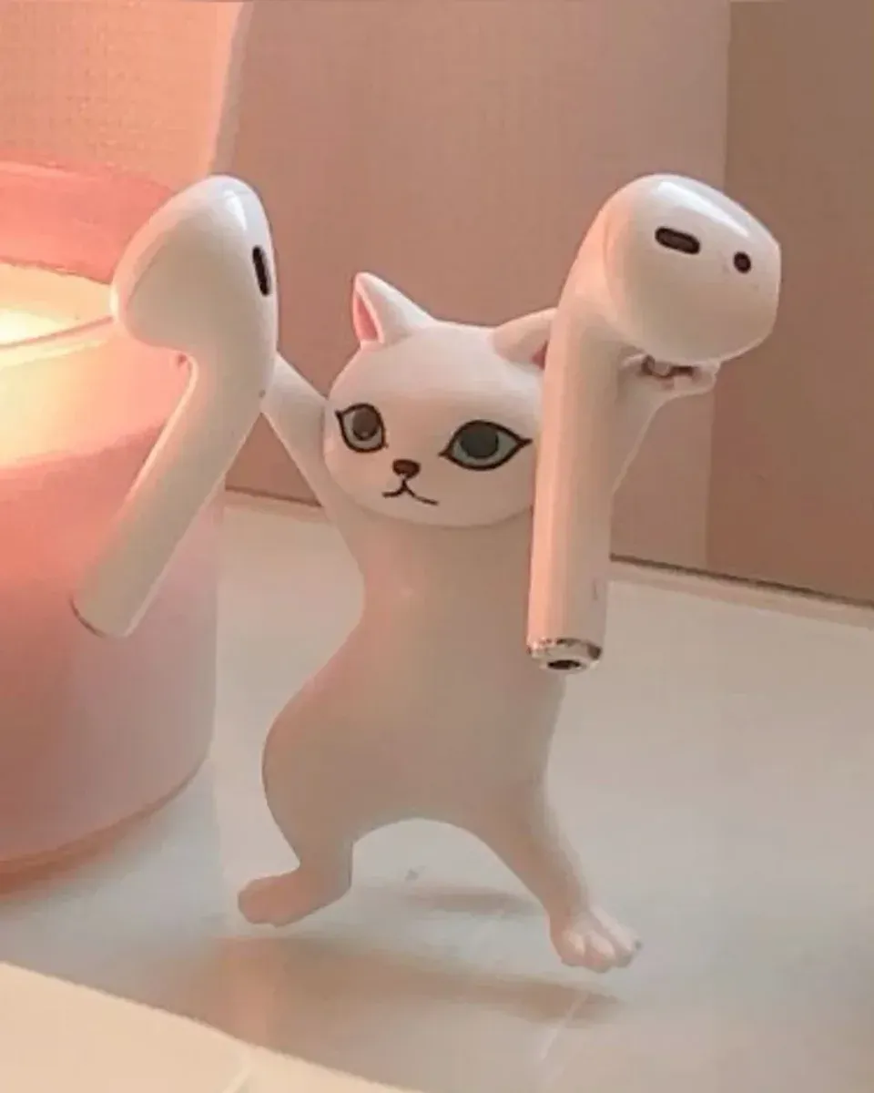 White cat figurine standing on its hind legs, balancing a single AirPod in each raised paw next to a pink candle