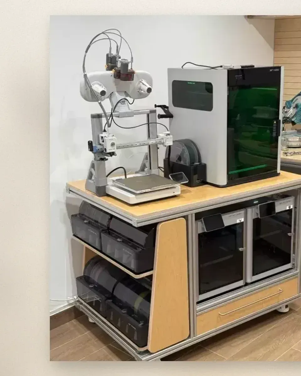 Open-frame printer on the top deck next to an enclosed printer with a lit chamber, fed by a wall-mounted filament dryer