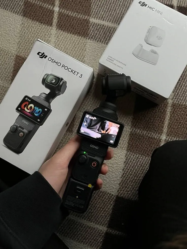 Hand holding the DJI Osmo Pocket 3 in front of its retail box and a DJI Mic Mini box